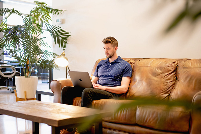 A man works diligently on his laptop from a brown leather couch in a modern, naturally-lit office. With a potted plant and lamp nearby, he focuses on conducting effective keyword research to guarantee SEO success.