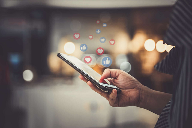 A person engages with their smartphone, showcasing heart and thumbs-up icons symbolizing their mastery of social media channels against a warmly lit, blurred background.