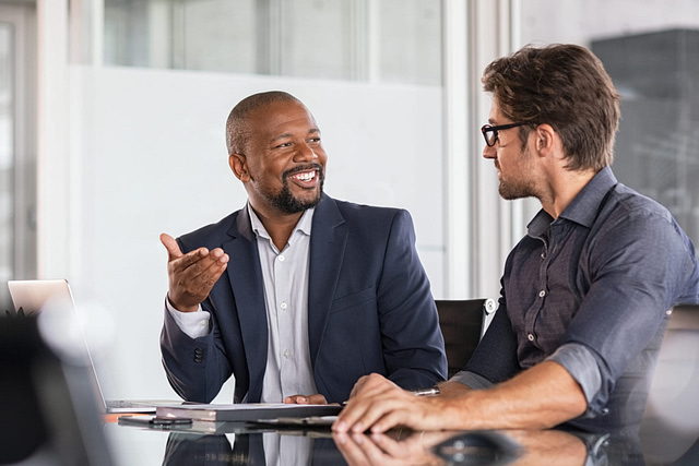In a modern office setting, a man in a navy blazer enthusiastically discusses strategies for acquiring more reviews, while another man in glasses and a gray shirt listens intently. A laptop sits open on the table between them.