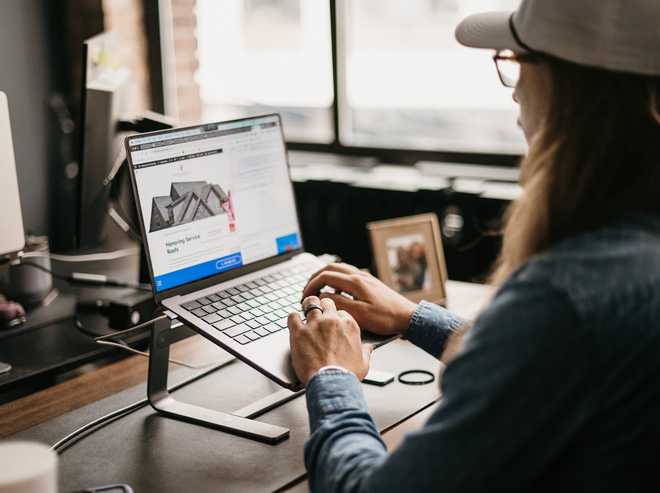 A long-haired individual wearing a cap sits at a desk using a laptop on a stand. The screen showcases Home Service PPC content. The workspace, featuring a framed photo and modern office chair, is bathed in natural light from a nearby window, fostering an inviting and productive environment.