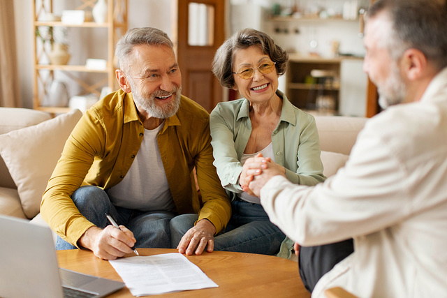 An elderly couple, seated cozily on their couch, warmly shakes hands with a representative across the table. Nearby, documents and a laptop indicate they are finalizing an agreement with an HVAC and plumbing financing company in their inviting living room.