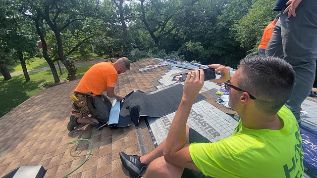 Two roofers on a shingled roof are actively engaged in their craft. While one expertly installs shingles, the other documents the process with a smartphone to enhance roofing SEO. Clad in casual work attire, they operate under a clear sky with trees providing a natural backdrop. Best roofing marketing companies