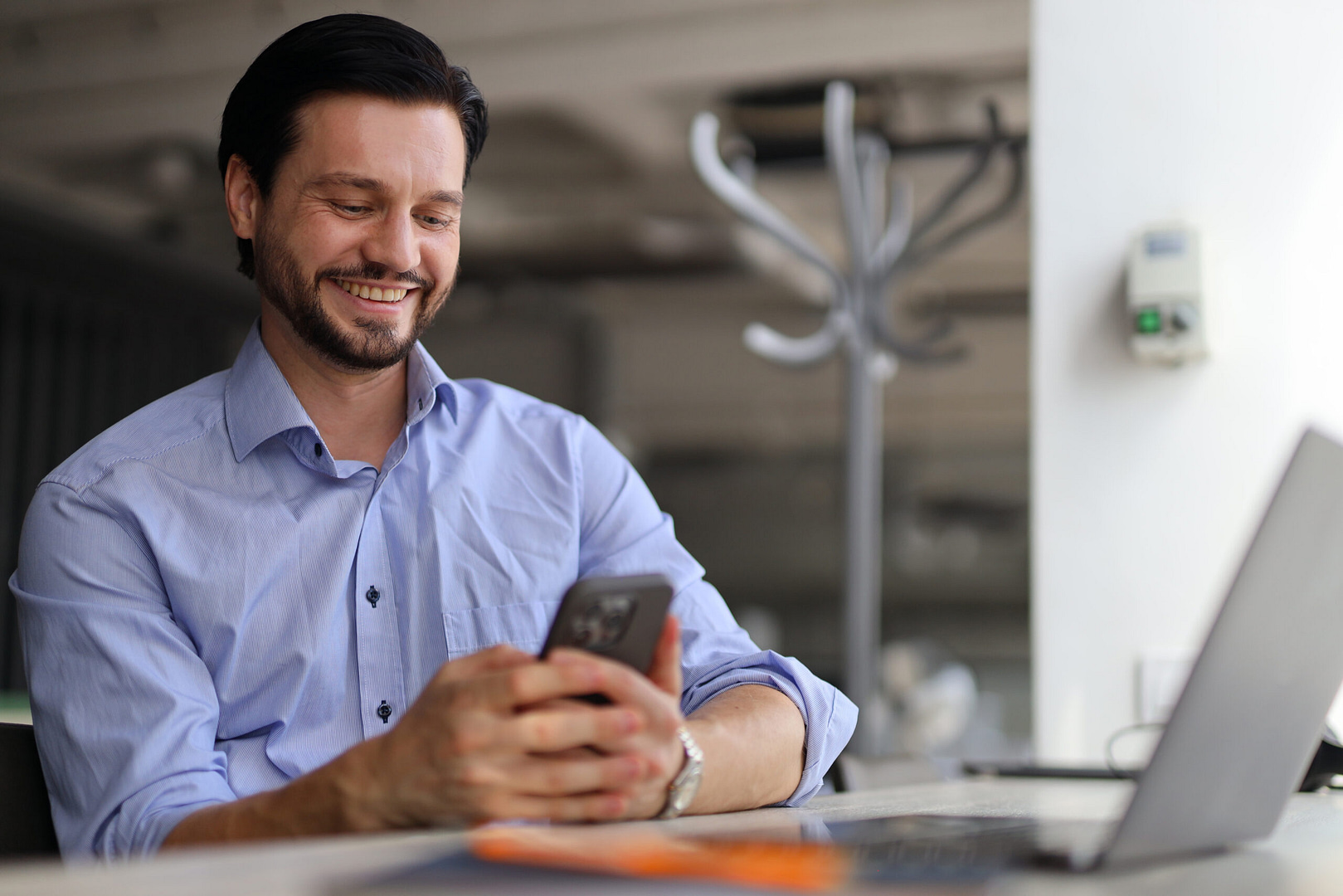 A man in a blue shirt smiles as he reviews Des Moines SEO updates on his phone, seated at his desk with a laptop in an office setting.