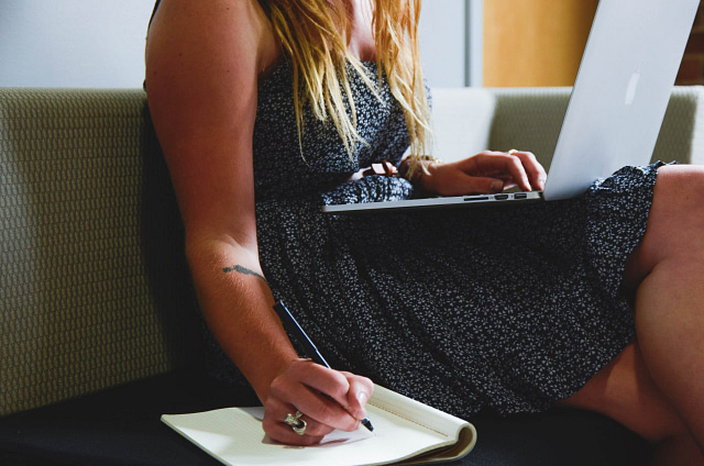 A person sits on a sofa with a laptop perched on their lap, wearing a black and white patterned dress. They scribble notes in a notebook with a pen in their right hand while showcasing a small tattoo on their left arm, capturing the essence of modern startups.