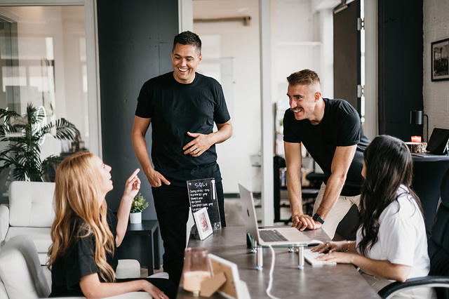 In an office setting at Hook Agency, four individuals engage in a dynamic conversation around a laptop on the table. With two standing and two seated, the scene reflects a collaborative atmosphere, underscoring the value of diverse perspectives.