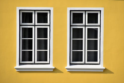 Consider replacing your windows with these identical, six-pane, white-framed options. Their sleek design enhances any space, as shown by the way they cast elegant shadows on a vibrant yellow wall. Ideal for those seeking both style and practicality in window replacements.