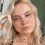 Courtney Owens, with long wavy blonde hair and a black top, faces the camera with a neutral expression. Layered necklaces adorn her as she stands outdoors, set against a soft-focus background.